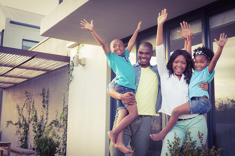 Client Center - Portrait of a Cheerful Family with Two Kids Standing Outside Their House with Their Hands Up in the Air